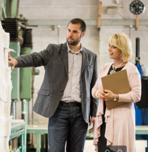 Man in a grey blazer showing fabric samples to a woman holding a clipboard inside a factory setting.